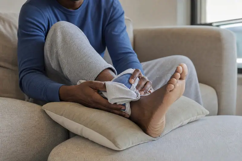 Personne assise appliquant une poche de glace sur sa cheville, jambe surélevée sur un coussin