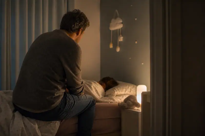 Parent assis au bord d’un lit d’enfant dans une chambre faiblement éclairée par une veilleuse