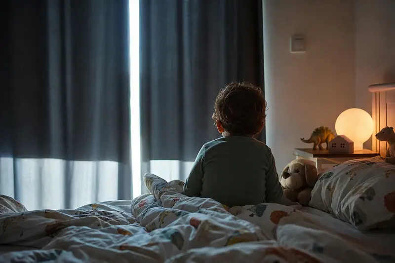Jeune enfant vu de dos, assis réveillé dans son lit à l’aube, lumière filtrant autour des rideaux