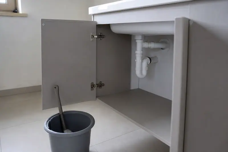 Meuble de salle de bain ouvert laissant voir le siphon en plastique blanc et les tuyaux d’évacuation sous le lavabo