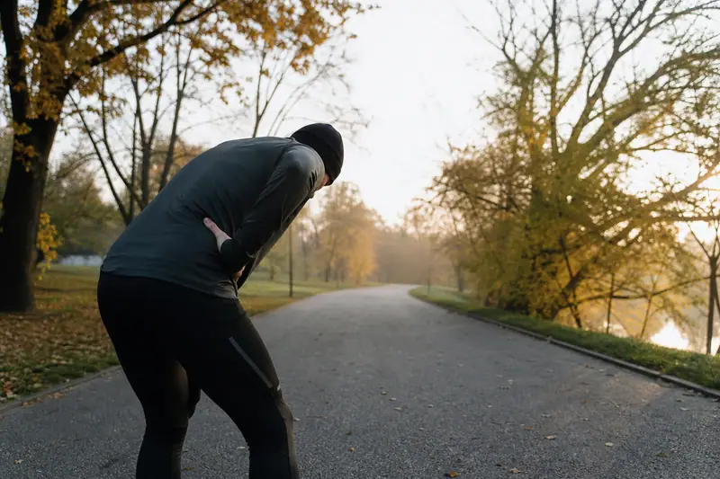 Coureur arrêté sur une allée de parc au petit matin, penché en avant, une main pressée sur les côtes pour reprendre son souffle
