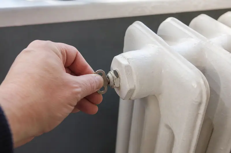 Radiateur en fonte blanc sous une fenêtre dans un salon, main tenant une clé de purge près de la vis en haut de l’appareil