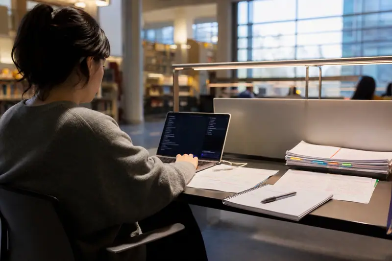 Étudiante utilisant un ordinateur et des fiches de révision dans une bibliothèque universitaire calme