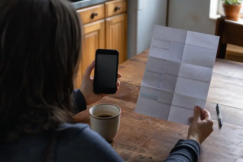 Personne attablée le matin, smartphone et relevé bancaire papier en main, l’air préoccupé