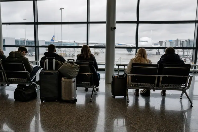 Passagers assis avec leurs valises dans un hall d’aéroport désert face aux baies vitrées, avion immobile sur le tarmac sous un ciel gris